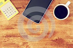 Top view of tablet, coffee cup and calculator over wooden textured table background.