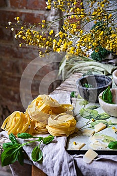 Top view of tagliatelle with basil, cheese and bowls on dishcloth, unfocused rustic background and mimosas