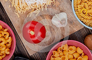 top view of tagliatelle macaroni with flour garlic and tomato on cutting board with other types of pasta on wooden background