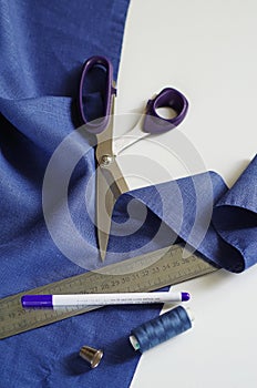 Top view tailor accessories and tools. Blank notepad surrounded with cutting scissors, spools of thread, violet fabric. Measuring