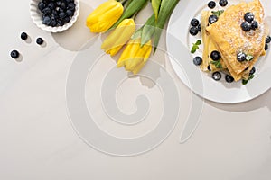 Top view of tasty crepes near bowl of blueberries and yellow tulips