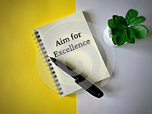 Top view of text on notebook - Aim for excellence. With pen and green plant background. Business concept