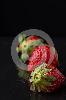 Top view of three strawberries, with selective focus, on wet black stone slate, black background, with reflection, vertical,