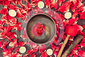 Top view tibetan singing bowl with floating inside in water red peony flower. Burning candles and petals on the black stone backgr