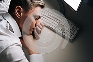 Top view of tired sleepy male employee asleep on computer desk late at night. Exhausted businessman sleeping with head