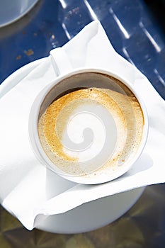 Top view to a hot fresh cup of capuccino in bright sunlight. White mug of hot coffee with milk on white napkin