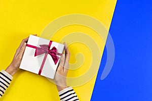 Top view to woman hands holding gift present box decorated with bow on yellow and blue background