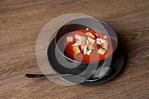 Top view of toothsome tomato soup with sour cream and croutons in black bowl on plate with spoon on brown wooden table.