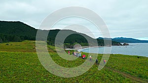 Top view of tourists hiking on green coast on cloudy day. Clip. Hikers walk along coastal path with green mountains and