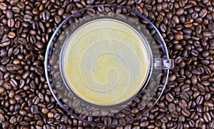 Top view on transparent glass cup, saucer and handle with black coffee whitecap crema, blurred roasted coffee beans background