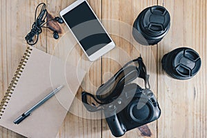 Top view of travel object : gray pencil on brown notebook. and white mobilephone, earphone, black camera and two lens on wooden ta