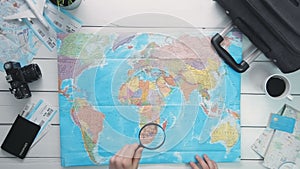 Top view traveler`s hands looking at world map using magnifying glass at white wooden desk