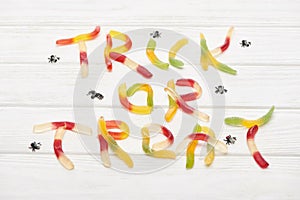 Top view of trick or treat lettering made of colorful gummy sweets on white wooden table with spiders, Halloween treat.