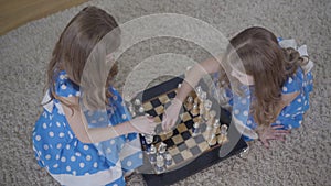 Top view of two brunette caucasian twin sisters playing chess indoors. Little girls in elegant blue dresses resting
