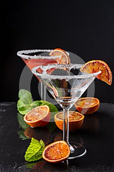Top view of two cocktail glasses with Blood Orange Martini, one empty, with selective focus, with half orange and mint, on black b