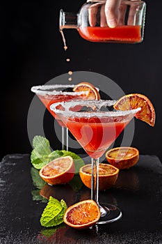 Top view of two cocktail glasses with unfocused bottle serving drops of blood orange martini, half orange and mint, on black backg