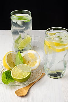 Top view of two glasses of water with lemon and lime slices with ice cubes on white wood, with wooden spoon, plate with half lemon