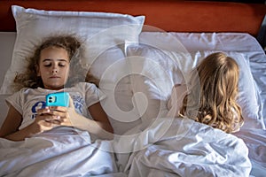 Top view of two little girls sisters playing with their smartphones lying on a bed in a large hotel room. harm of