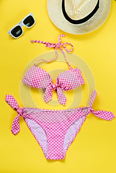 Top view of two pieces pink chequered swimming suit, white sunglasses and straw hat over yellow background.