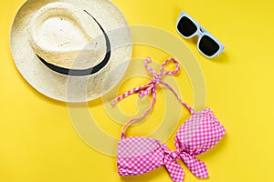 Top view of two pieces pink chequered swimming suit, white sunglasses and straw hat over yellow background.