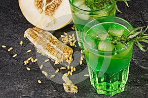 Top view of two refreshing green cocktails with tarragon leaves and cubes of ice on a black background. A big juicy melon with see