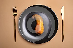 Top view of ugly bent ripe pear lies on round black plate with golden cutlery on beige background.