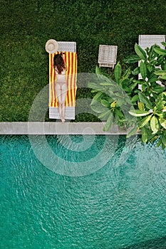 Top view of unrecognizable slim young woman in beige bikini relaxing and sunbathe near luxury swimming pool in green tropic in