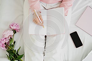 Top view of unrecognizable woman sitting on bed and making notes to her diary.