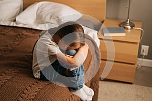 Top view of upset little girl hugging knee, sobbing with head bowed and crying sitting alone on bed in bedroom.