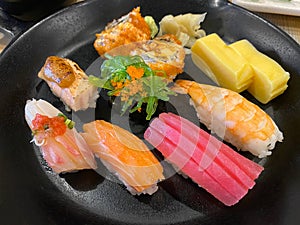 Top view of variety of sushi and rolls served on black plate. Plenty of nigiri, maki, and roll with tuna, salmon, shrimp