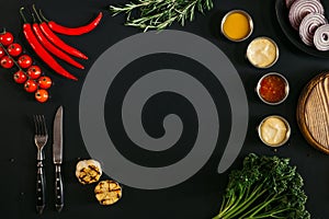 top view of various sauces, grilled garlic, fork with knife and fresh vegetables with herbs on black background
