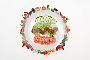 Top view of vegetables arranged in round frame around falafel on plate on white background.