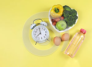 Vegetables  and fruit  in heart shape plate  with white vintage alarm clock, eggs  and pink bottle of water on yellow background.