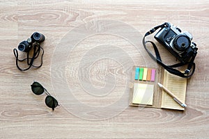 Top view vintage camera, sunglasses, binoculars and notepad with pen on the wooden background with copy space for adding some text