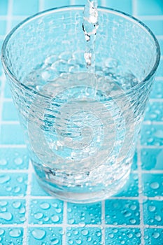 Top view of water jet falling into a glass cup, with selective focus, on wet light blue mosic, in vertical,