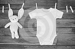 Top view white baby bodysuit on a clothesline with teddy hare on a wooden background. Copy space for text mockup