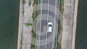 Top view of white car rides through dam route. SUV drives at road near lake on summer day. Flying over the auto moves