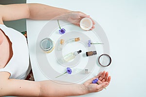 Top view of white caucasian woman sitting at white table with cosmetical flasks and jars in front of her, a cream jar and a blue