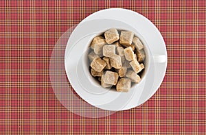 Top view of white coffee or tea cup full brown sugar cubes on red squared background. Diet unhealty sweet addiction