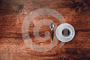 Top view of white Cup with black coffee on a white saucer and teaspoon on dark brown wooden background