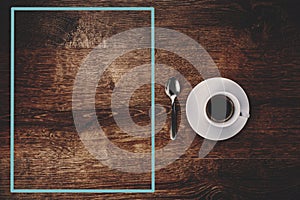 Top view of white Cup with black coffee on a white saucer and teaspoon on dark brown wooden background with blue frame on the left