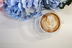 Top view of white cup of hot coffee latte with milk foam heart shape art on white table with blue and purple hydrangea flower