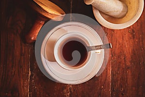 Top view of white cup and a saucer filled with herbal tea and metal spoon, stone mortar and a pestle on a wooden background.