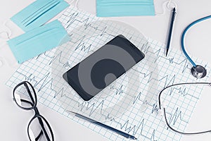 Top view of white desktop with empty cellphone, stethoscope, cardiogram, pen, other items and mock up place. Doctor`s office,