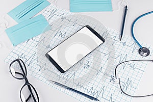 Top view of white desktop with empty smartphone, stethoscope, cardiogram, pen, other items and mock up place. Doctor`s office,