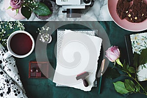 Top view of white paper sheets, coffee, rose, stamps, cake, camera on a green background