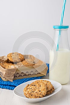 Top view of white plate with chocolate chip cookies, bottle of milk with blue straw, metal box with cookies and blue wool cloth, o