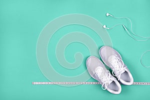 Top view of white sport shoes sneakers with measuring tape and white earphones headphones on blue-green background.