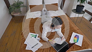 Top view wide shot of young Asian man lying on bed in headphones and Covid-19 face mask messaging online. Male Chinese