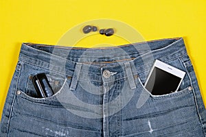 Top view of wireless headphones, jeans, charger and smartphone in its pocket on yellow background